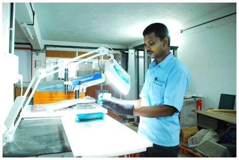 Technician inspecting a sheet metal part under an illuminated magnifier at the finishing bench